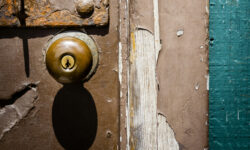 Close-up of a tarnished doorknob set in a door covered in peeling paint, and bits of rusted metal. It is an old and weathered door.