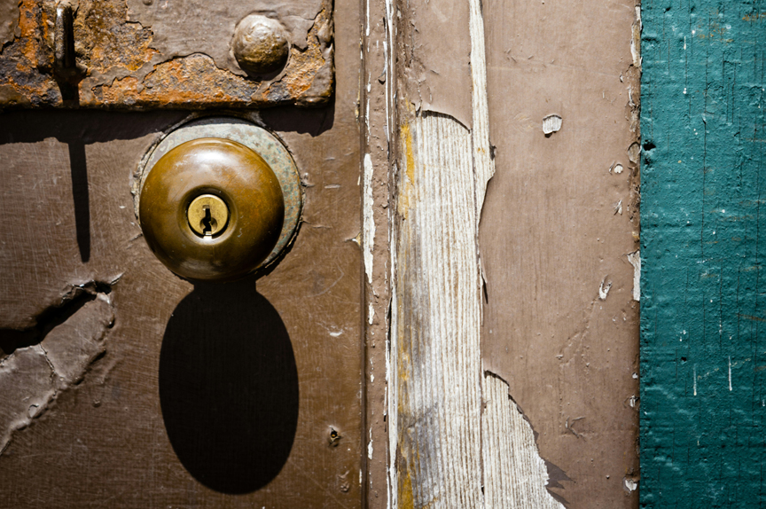 Close-up of a tarnished doorknob set in a door covered in peeling paint, and bits of rusted metal. It is an old and weathered door.